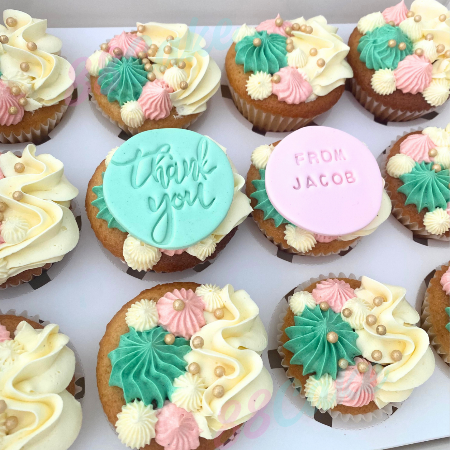 A close-up of several decorated cupcakes arranged on a white surface. The cupcakes have cream-colored frosting topped with mint green and pastel pink piped swirls, small pearl-like beads, and two round topper pieces: one light aqua with the words 'thank you' and one light pink with the words 'FROM JACOB'.