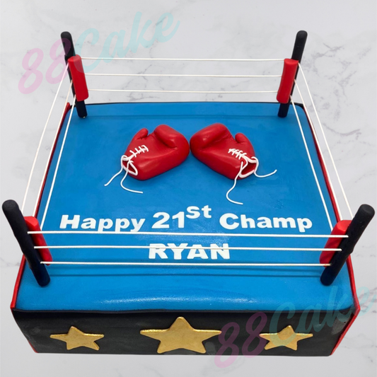 Cake designed like a boxing ring with red boxing gloves and text 'Happy 21st Champ Ryan' on a light gray background.